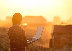 Young woman with laptop standing on field in sunset and looking at tractor baling